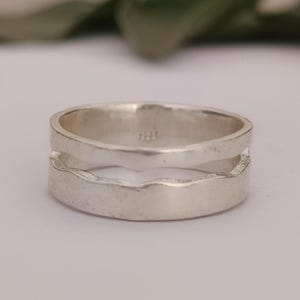 Puede incluir: Un anillo de plata con un diseño único. El anillo presenta una banda dividida con un patrón ondulado y texturizado. El metal tiene un acabado mate y el anillo parece ser de plata de ley. El anillo es una joya.