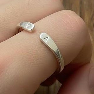 Puede incluir: Un anillo abierto de color plateado con las letras "A" y "V" grabadas en cada extremo. El anillo se lleva en un dedo, mostrando su diseño minimalista y su acabado pulido.