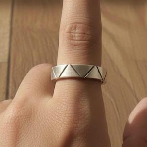 May include: A silver ring with a geometric triangle design. The ring is worn on a finger, showcasing its modern and minimalist style. The ring's design features a series of repeating triangles around the band, creating a unique and eye-catching pattern.
