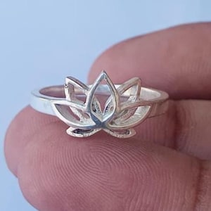 Può includere: Un anello d'argento con un design a fiore di loto. L'anello ha una fascia sottile e il fiore di loto è il punto focale, con dettagli intricati dei petali. L'anello è mostrato su un dito, evidenziando la delicata maestria e il design.