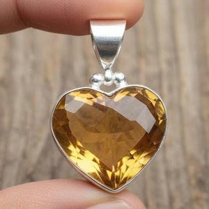 May include: A heart-shaped pendant with a faceted, golden-yellow gemstone set in a silver metal frame. The pendant is held up by a silver bail with two small decorative spheres. The gemstone has a brilliant cut.