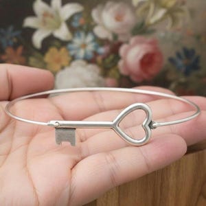 Può includere: Un braccialetto a cerchio d'argento con un ciondolo a forma di chiave, la cui testa ha la forma di un cuore. Il ciondolo a forma di chiave è attaccato al cinturino del braccialetto. Il braccialetto è tenuto in mano, con uno sfondo floreale sfocato.