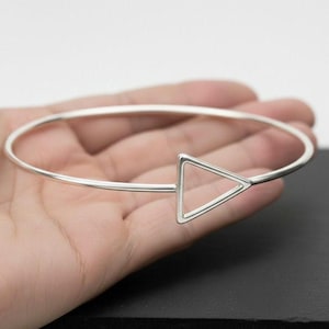 May include: A silver bangle bracelet with a geometric triangle design. The bracelet is a simple, minimalist style, with a thin, circular band and a hollow triangle accent. The jewelry is shown on a hand.