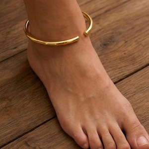 May include: A polished gold-tone metal anklet featuring a simple, open-ended circular band design. The jewelry piece has a smooth, reflective surface and a minimalist, continuous hoop structure that rests around the ankle area for a clean look.