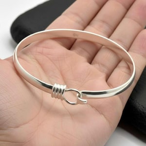 May include: A silver bangle bracelet with a hook and eye closure. The bracelet is round and has a smooth, polished finish. The hook and eye closure is a simple design, with a wrapped wire detail. The bracelet is shown on a person's hand.