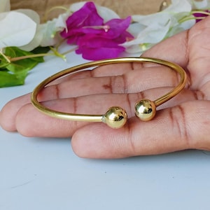 May include: A gold-colored bangle bracelet with two spherical end caps. The bracelet is open-ended and appears to be made of a shiny metal. The background includes green leaves and pink and white flowers.