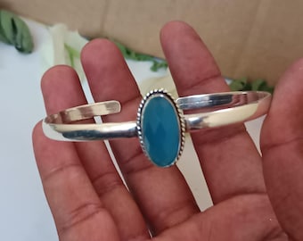 Könnte beinhalten: Ein silberfarbenes Armreif-Armband mit einem ovalen, hellblauen Edelstein. Der Edelstein ist in einer Perlenfassung gefasst. Das offene Armband ermöglicht eine einfache Anpassung. Das Armband wird in einer Hand gehalten.