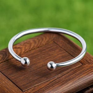 May include: A polished silver open cuff bracelet. The bracelet has a rounded design with a ball at each end. The jewellery is displayed on a wooden surface with a green background. The bracelet's diameter is approximately 6 cm.