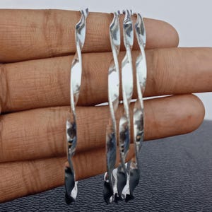 May include: Four silver-coloured, twisted metal bangles. Each bangle has a spiral design, reflecting light. The bangles are displayed against a neutral background, showcasing their reflective quality and design. These bracelets are a simple yet elegant accessory.