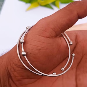 Puede incluir: Dos pulseras plateadas con un diseño minimalista. Cada pulsera presenta una banda fina y curvada con pequeños acentos esféricos. Las pulseras se muestran sobre un fondo color piel, resaltando su estética elegante y moderna. Joyería.