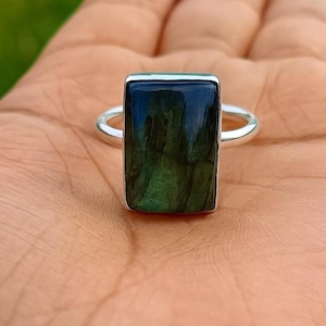 May include: A rectangular gemstone ring with a silver band. The gemstone is a dark blue-green color with a rectangular shape. The ring is set in a silver band.