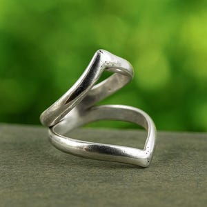 Może przedstawiać: Srebrny pierścionek o unikalnym, otwartym designie. Pierścionek posiada dwa zakrzywione paski, które się przecinają, tworząc kształt litery V. Metal ma polerowane wykończenie, odbijające światło. Pierścionek jest umieszczony na rozmytym zielonym tle.