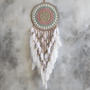 May include: A decorative dreamcatcher with a circular design. The centre features a detailed, multi-coloured web with pink, green, and beige accents. White feathers and small beads hang from the bottom, creating a bohemian aesthetic. It hangs against a grey background.