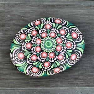 May include: A hand-painted, oval-shaped stone featuring a mandala design. The design includes a central green circle, surrounded by coral-colored circles, white dots, and green leaf-like shapes on a black background. The stone is approximately 5 inches long.