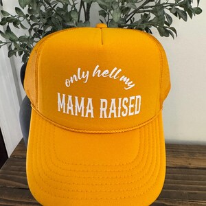 May include: Mustard yellow trucker hat with the text "only hell my MAMA RAISED" in white block letters. The hat has a mesh back and a curved brim. A fun and bold accessory.