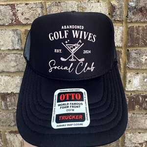 Op de afbeelding: Zwarte truckerpet met witte tekst "ABANDONED GOLF WIVES SOCIAL CLUB EST. 2024". De pet heeft een mesh achterkant en een gebogen klep. Een witte sticker op de voorkant leest "OTTO WORLD FAMOUS FOAM FRONT TRUCKER".