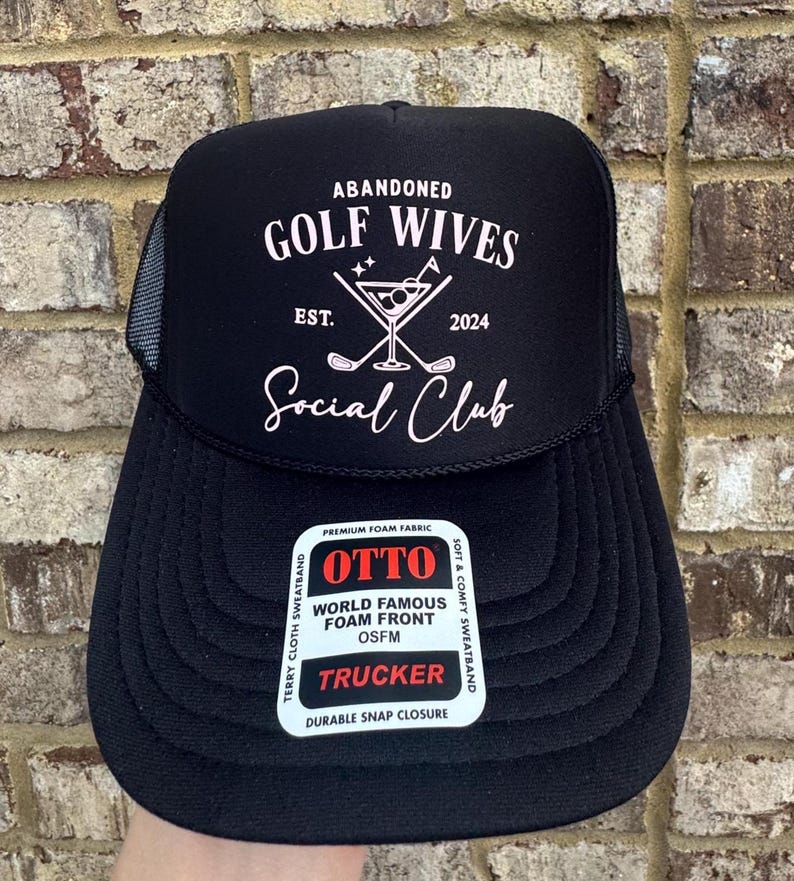 Op de afbeelding: Zwarte truckerpet met witte tekst "ABANDONED GOLF WIVES SOCIAL CLUB EST. 2024". De pet heeft een mesh achterkant en een gebogen klep. Een witte sticker op de voorkant leest "OTTO WORLD FAMOUS FOAM FRONT TRUCKER".