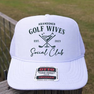 Op de afbeelding: Witte truckerpet met een mesh achterkant en een gebogen klep. De pet heeft de tekst "ABANDONED GOLF WIVES Social Club" in donkergroen, samen met een cocktail en golfclub afbeelding. De pet is gemaakt van schuim en heeft een druksluiting.