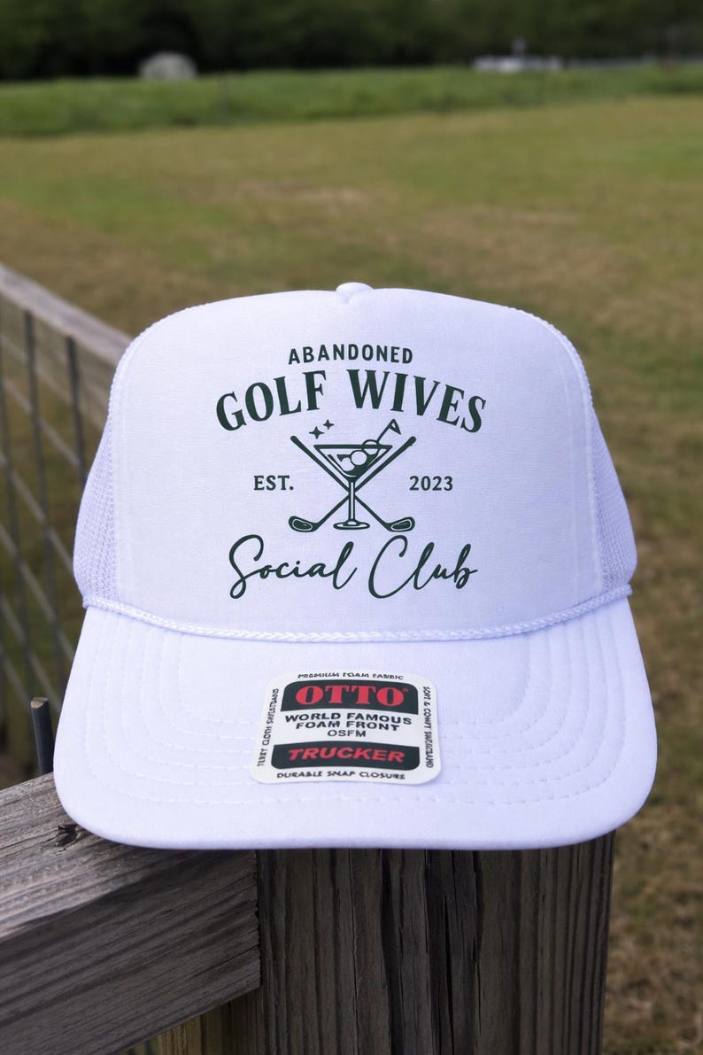 Op de afbeelding: Witte truckerpet met een mesh achterkant en een gebogen klep. De pet heeft de tekst "ABANDONED GOLF WIVES Social Club" in donkergroen, samen met een cocktail en golfclub afbeelding. De pet is gemaakt van schuim en heeft een druksluiting.