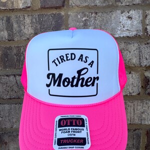 K&ouml;nnte beinhalten: Neonpinke und wei&szlig;e Trucker-Cap mit dem Text "TIRED AS A Mother" in schwarzer Schrift. Die Kappe hat eine Schaumstofffront und ein Mesh-R&uuml;ckenteil mit einem Schnappverschluss. OSFM (One Size Fits Most).