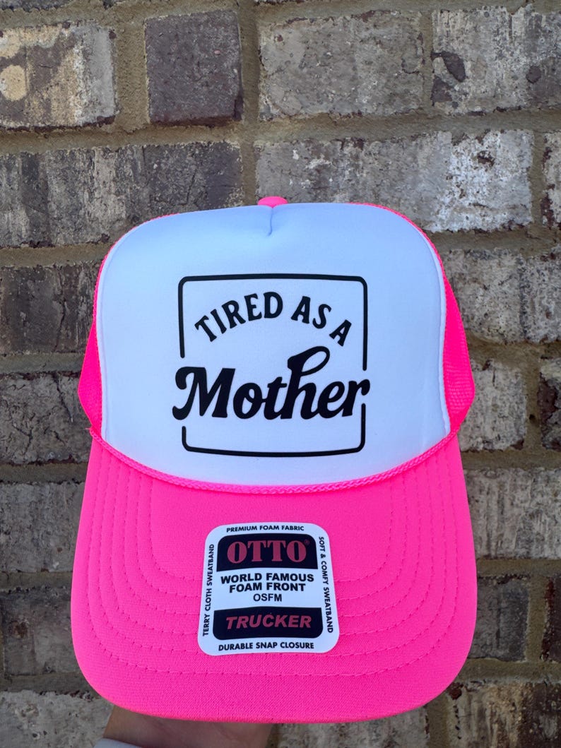 K&ouml;nnte beinhalten: Neonpinke und wei&szlig;e Trucker-Cap mit dem Text "TIRED AS A Mother" in schwarzer Schrift. Die Kappe hat eine Schaumstofffront und ein Mesh-R&uuml;ckenteil mit einem Schnappverschluss. OSFM (One Size Fits Most).