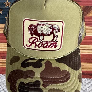 May include: A green and brown camouflage trucker hat with a patch featuring a bison and the word "Roam" in burgundy. The hat has a mesh back and a curved brim.