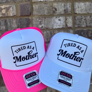 K&ouml;nnte beinhalten: Zwei Trucker-H&uuml;te mit dem Text "Tired as a Mother" in schwarzer Schrift auf einer wei&szlig;en Frontplatte. Einer hat einen leuchtend rosa Rand und der andere einen wei&szlig;en Rand. Beide haben einen wei&szlig;en Mesh-R&uuml;cken.
