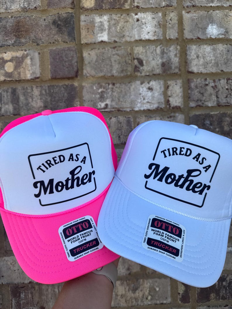 K&ouml;nnte beinhalten: Zwei Trucker-H&uuml;te mit dem Text "Tired as a Mother" in schwarzer Schrift auf einer wei&szlig;en Frontplatte. Einer hat einen leuchtend rosa Rand und der andere einen wei&szlig;en Rand. Beide haben einen wei&szlig;en Mesh-R&uuml;cken.