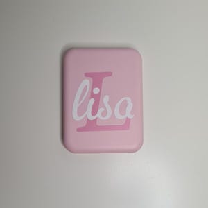 May include: A rectangular, pale pink object with rounded corners. The word "Lisa" is written in white cursive script over a pink "L" initial. The object is likely a phone case or similar accessory.