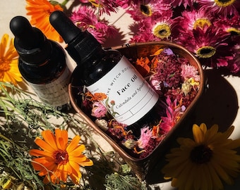 Puede incluir: Dos botellas negras con cuentagotas de aceite facial con etiquetas blancas, rodeadas de flores naranjas y rosas. Una botella está etiquetada como "Face Oil Calendula and Spilanthes". La escena se desarrolla sobre una superficie clara con hierbas secas.