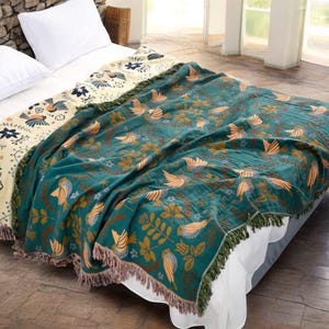 May include: A teal throw blanket with a bird and floral pattern, fringed edges, and a cream-coloured border. The blanket is draped over a bed with white pillows and a white bedspread. The blanket's design includes orange and brown birds and leaves.