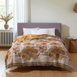 Könnte beinhalten: Eine floral gemusterte Decke mit Fransen ist über ein Bett drapiert. Die Decke zeigt große, stilisierte Blumen in Gold-, Braun- und Cremetönen. Das Bett hat weiße Kissen und ein graues Kopfteil. Die Decke misst ca. 178 cm x 203 cm.