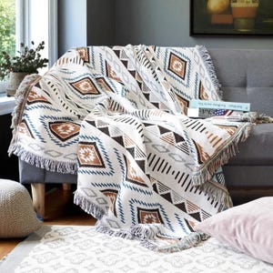 Puede incluir: Una manta tejida de color crema con un patrón geométrico de diamantes y triángulos marrones, azules y grises. La manta tiene un borde con flecos y está colocada sobre un sofá gris. Una alfombra a juego es visible en el suelo.