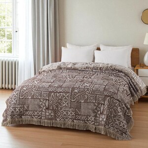 Könnte beinhalten: Eine braun-weiß gemusterte Wurkdecke mit Fransen ist über ein Bett drapiert. Die Decke weist ein sich wiederholendes geometrisches und florales Design auf. Das Bett hat weiße Kissen und ein hölzernes Kopfteil. Der Raum hat ein Fenster mit Vorhängen.