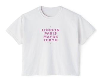 May include: White t-shirt with a crew neck and short sleeves. The shirt features the text "LONDON PARIS MAYBE TOKYO" in a lavender color, centered on the chest. The design includes small star-like accents.
