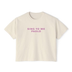 May include: A cream-coloured t-shirt featuring the lavender text "SING TO ME PAOLO". The text is centrally placed. The shirt has a crew neck and short sleeves.