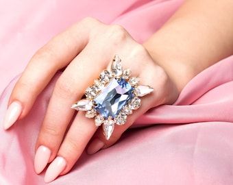 May include: A large, blue, rectangular gemstone ring with a gold setting and smaller clear gemstones surrounding the center stone. The ring is worn on a hand with pink nail polish.