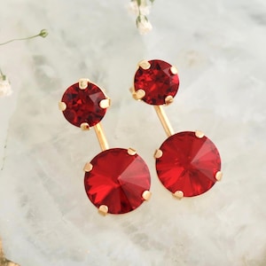 May include: A pair of red crystal earrings with gold-colored posts and settings. The earrings feature a large, round, faceted crystal at the bottom and a smaller crystal at the top. The earrings are embellished with Swarovski crystals.