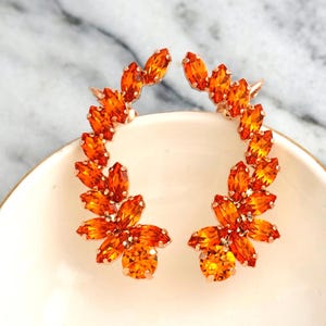 May include: A pair of orange crystal ear cuffs in a floral design. The ear cuffs are embellished with Swarovski crystals and are displayed on a white dish against a marble background. The text "ILONA RUBIN petite delights" is visible.