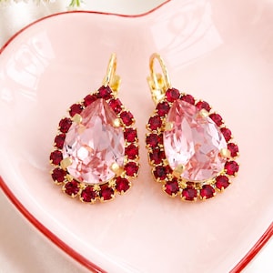 May include: A pair of gold-tone leverback earrings featuring a large, central pear-shaped light pink crystal surrounded by a halo of smaller, vibrant red round crystals. The jewelry is displayed on a heart-shaped dish with the text Ilona Rubin.
