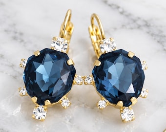 May include: A pair of gold-tone leverback earrings featuring a large, faceted, dark blue, octagonal crystal surrounded by six smaller, clear, round crystals. The text "ILONA RUBIN" and "Atelier-made jewelry" is printed at the bottom.
