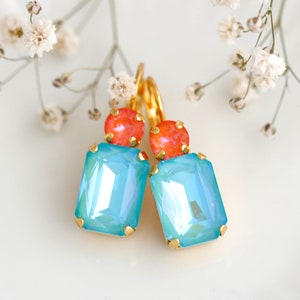 May include: A pair of drop earrings featuring a rectangular light blue crystal set in gold-tone metal, topped with a smaller round orange crystal. The earrings have a lever-back closure and are displayed with small white flowers.
