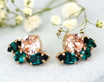 May include: A pair of gold-toned stud earrings. Each earring features a large, square, peach-colored crystal surrounded by smaller, emerald-green crystals. The earrings are set against a soft, light blue background, with delicate white flowers in the background.