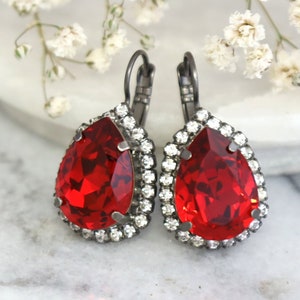 Op de afbeelding: Een paar oorhangers met een traanvormige rode kristal omgeven door een halo van kleinere heldere kristallen. De oorbellen hebben een zwarte metalen setting.