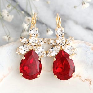 May include: A pair of gold-tone earrings featuring a large, pear-shaped red crystal drop suspended beneath a cluster of clear, marquise-cut crystals. The earrings have a lever-back closure and are branded with the text ILONA RUBIN petite delights.