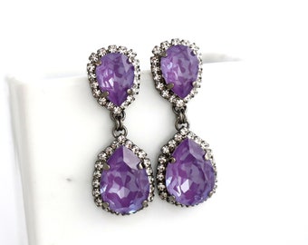 May include: A pair of drop earrings featuring large, pear-shaped purple crystals. Each crystal is surrounded by a halo of small, clear crystals. The earrings are suspended from a smaller, matching crystal. The metal has a dark, gunmetal finish.