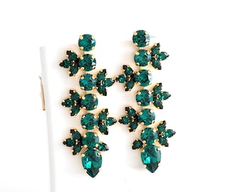 May include: A pair of gold-tone drop earrings featuring deep emerald green faceted crystals arranged in a floral-inspired cluster pattern. Each earring consists of multiple round and marquise-shaped stones set in metal prongs, creating a vertical design.