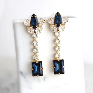 Puede incluir: Un par de pendientes largos y colgantes con un racimo de cristales transparentes y azul oscuro en la parte superior y un solo cristal azul oscuro rectangular en la parte inferior.