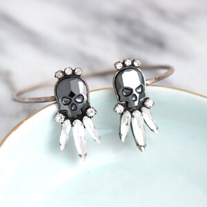 May include: A pair of silver skull earrings with crystal accents. The skulls are black and the crystals are clear.