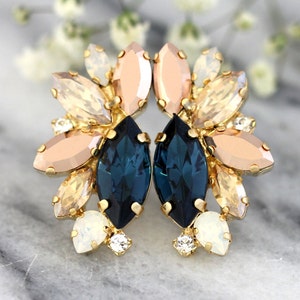 May include: A pair of stud earrings featuring a cluster of marquise-cut crystals in deep navy blue, soft peach, and iridescent white, all set in a gold-toned metal frame. The arrangement creates a floral-inspired shape with layered stones.